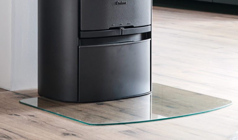 Svart kamin på trägolv, placerad på en skyddande glasplatta. Kaminen är enkel och modern i design.