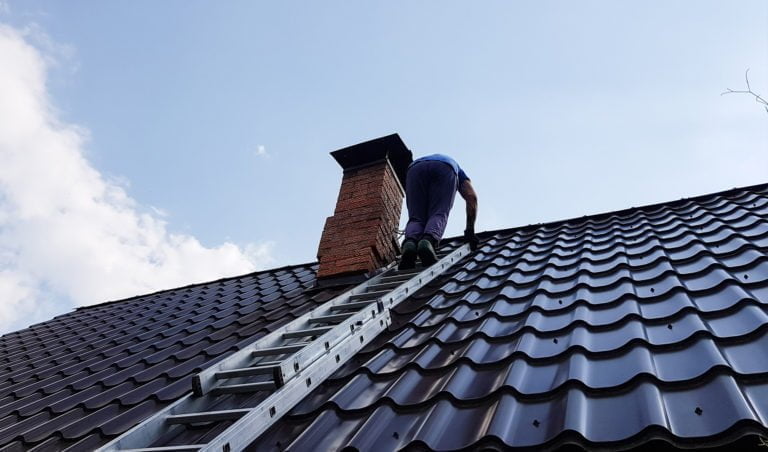 Person klättrar på en stege uppför ett plåttak bredvid en skorsten under klarblå himmel.