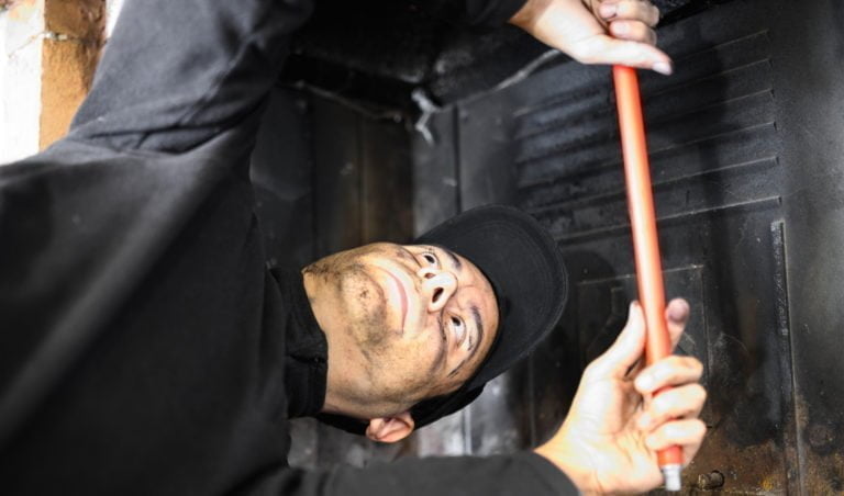 Person rengör insidan av en eldstad med ett rött verktyg, iförd mörka arbetskläder och keps. Eldstadsunderhåll.