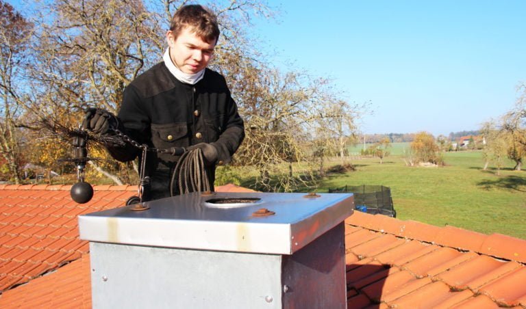 Sotare rengör skorsten på rött tegeltak med verktyg, omgiven av landskap med träd och gräs under klarblå himmel.