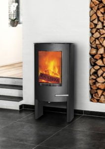 En modern, svart kamin med brinnande eld, står på ett klinkergolv bredvid en stapel vedträn och en vit vägg.