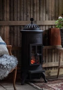 Svart gjutjärnskamin med glaslucka och synliga lågor, placerad i ett mysigt rum med träpaneler och fåtölj med fårskinn.
