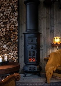 Svart kamin med vedstapel och tända ljus i mysig rustik miljö. Vedträ staplade till vänster, guldfärgad lampa och filt på fåtölj till höger.
