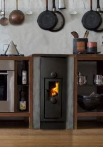 Kök med vedeldad spis, omgiven av köksredskap och hyllor med flaskor. Elden brinner inuti spisen, vilket skapar en varm atmosfär.