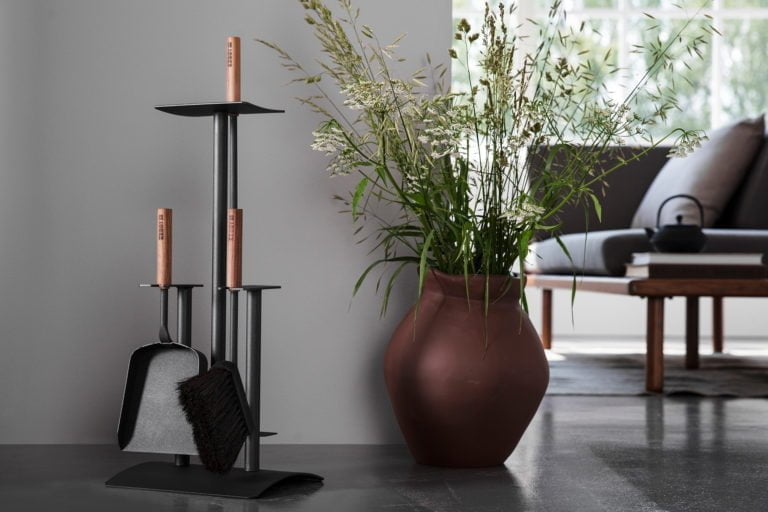 Eldstädsverktyg med trähandtag bredvid en brun vas med gröna växter i ett modernt vardagsrum.