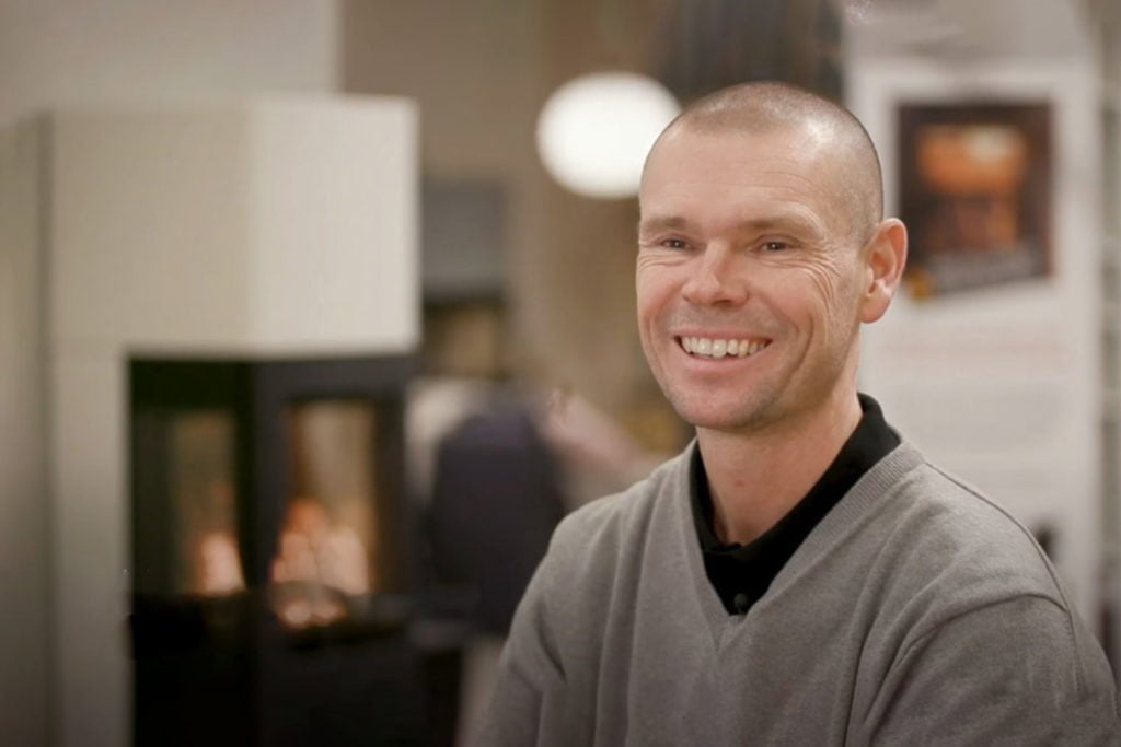 En leende man i grå tröja står framför en eldstad i en butiksmiljö. Elden brinner i bakgrunden, vilket skapar en varm atmosfär.