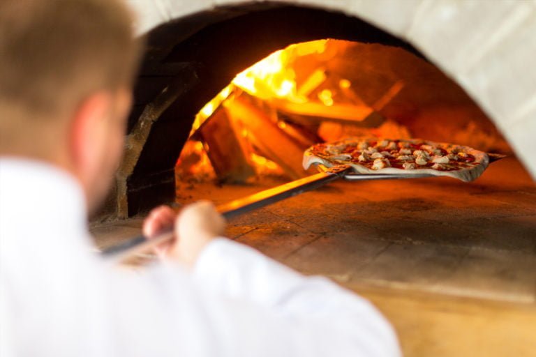 En pizza gräddas i en vedeldad ugn med eld i bakgrunden, hanterad av en person i vit skjorta.