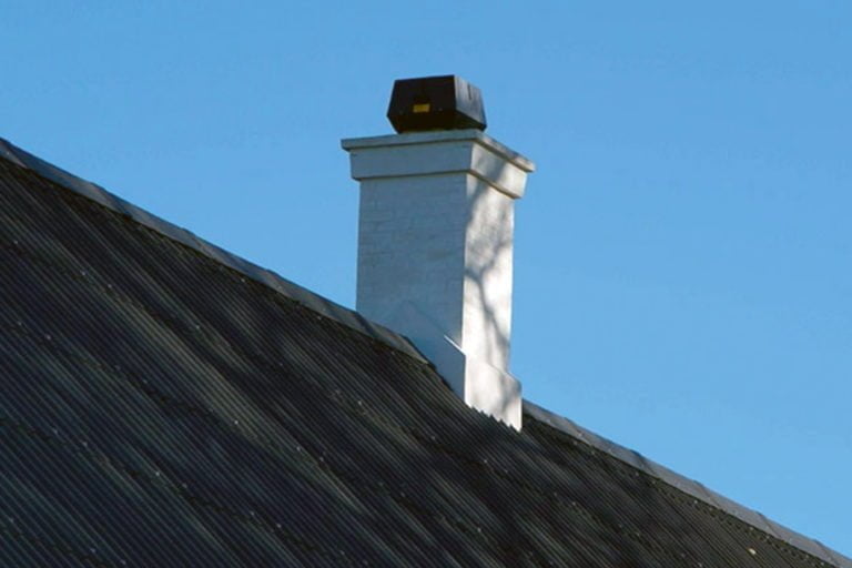 Vit skorsten med svart skorstenshuv på ett svart plåttak mot klarblå himmel.