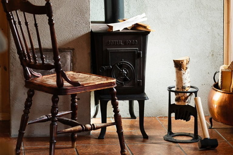 En gammal trästol och en vedkamin märkt "Jøtul 1940" står på ett klinkergolv, omgiven av ved och en yxa i ett hemtrevligt rum.
