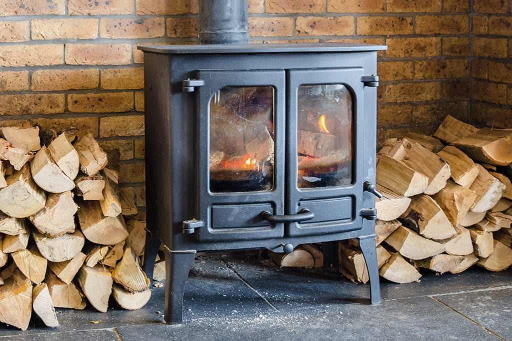 Svart vedkamin med brinnande eld, omgiven av staplat ved, mot en tegelvägg i ett rum med stengolv.