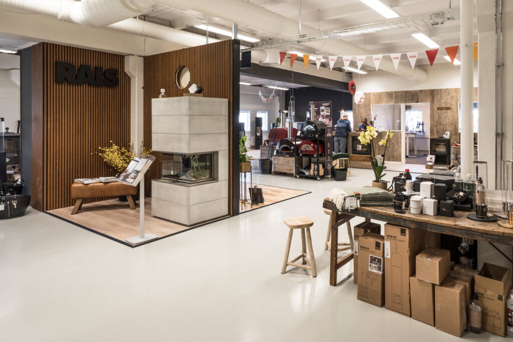 Interiör från en butik med utställning av moderna kaminer och tillbehör. Eleganta möbler, dekorationer och förpackade produkter syns i ljus och stilren miljö.