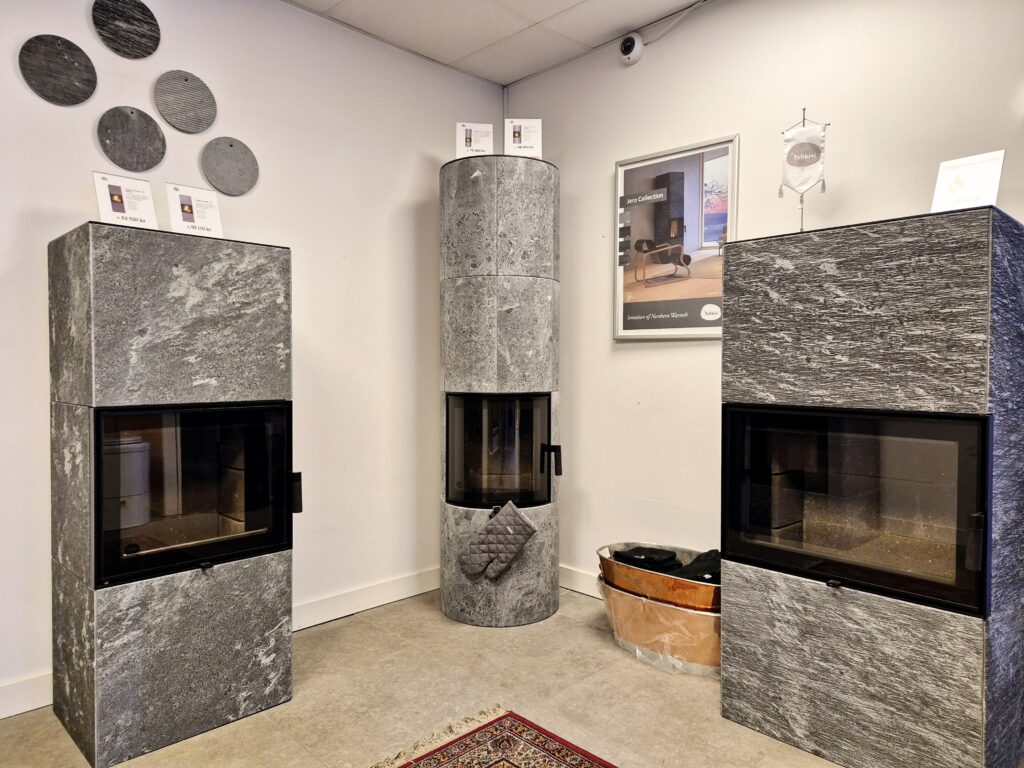 Tre stilrena kaminer i grå sten pryder ett showroom. En rund kamin i mitten, två rektangulära på sidorna. Dekorativa stenar på väggen ovanför.