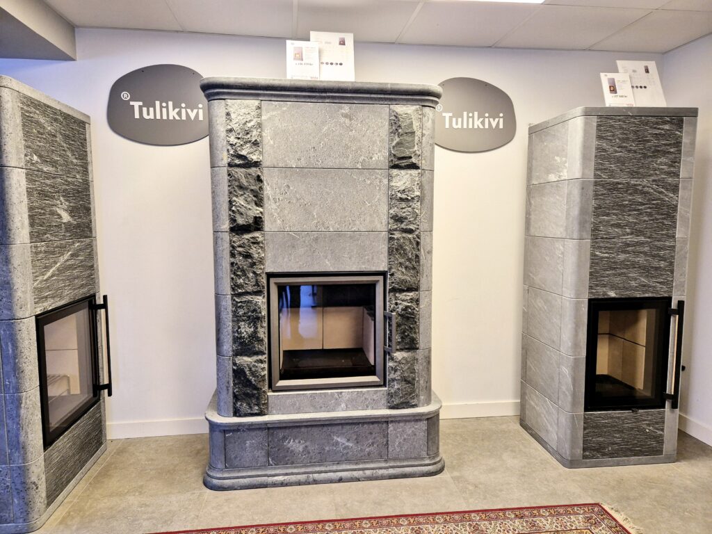 Tre gråa Tulikivi-kaminer i sten visas i ett showroom med skyltar ovanför. Kaminerna står på ett ljusgrått golv med en del av en röd matta synlig.