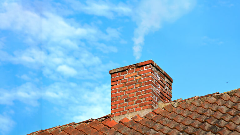 Tegelstensskorsten med rök, belägen på ett tak med tegelpannor, mot en klarblå himmel med moln.